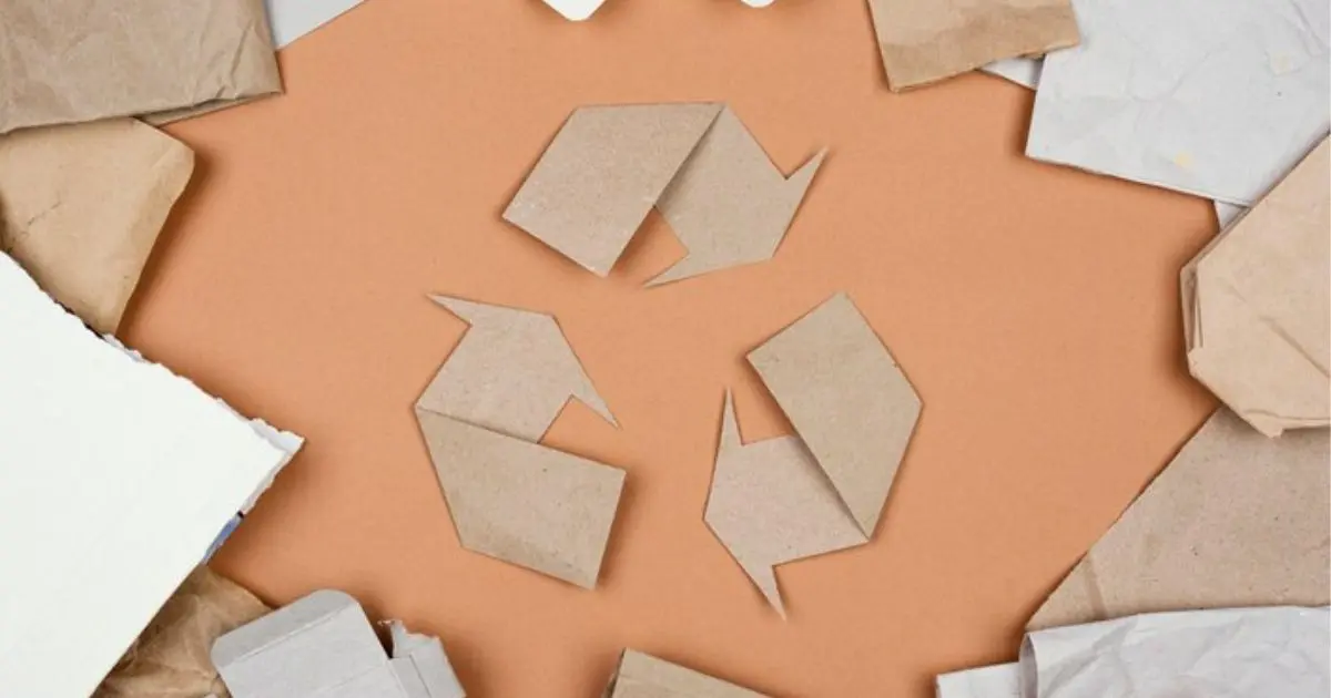 Símbolo de reciclagem feito de papelão, cercado por pedaços de papel reciclável em fundo laranja claro.