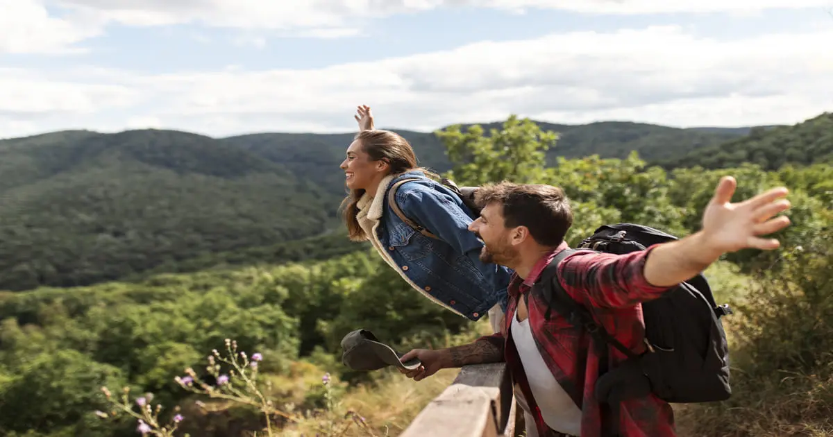 Casal sorridente em viagem sustentável, apreciando a vista de montanhas e floresta, com os braços abertos em sinal de liberdade.