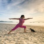 Mulher fazendo pose de yoga na praia ao nascer do sol, com um cachorro sentado ao lado observando a prática.