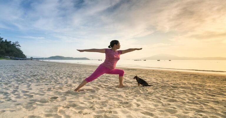 Mulher fazendo pose de yoga na praia ao nascer do sol, com um cachorro sentado ao lado observando a prática.