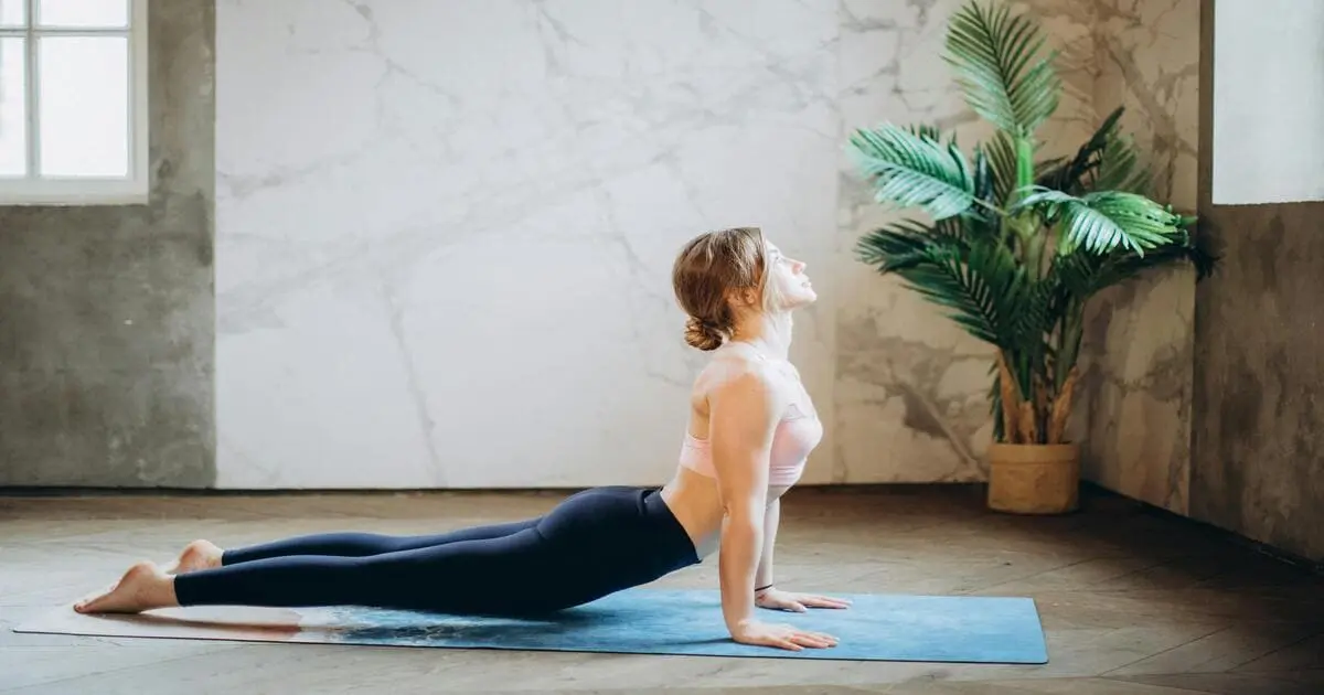 Mulher praticando yoga para iniciantes em casa na postura da cobra, deitada sobre um tapete com uma planta ao fundo em ambiente tranquilo.