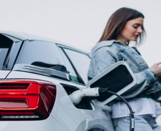 Mulher de jaqueta jeans usa o celular enquanto seu carro elétrico carrega na estação. Combinando modernidade e sustentabilidade.