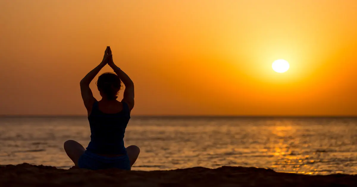 Pessoa meditando na praia ao nascer do sol, mãos unidas acima da cabeça, expressando calma, equilíbrio e conexão com a natureza na dinacharya.