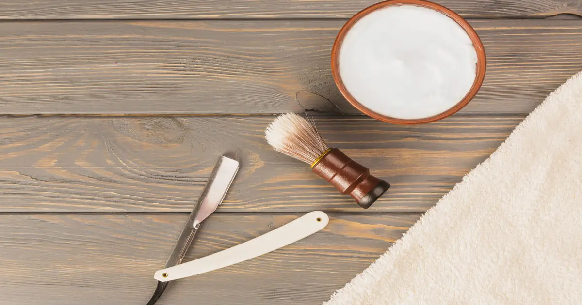 Kit de barbear clássico com navalha, pincel e creme de barbear caseiro sobre madeira, ao lado de uma toalha para um barbear confortável.