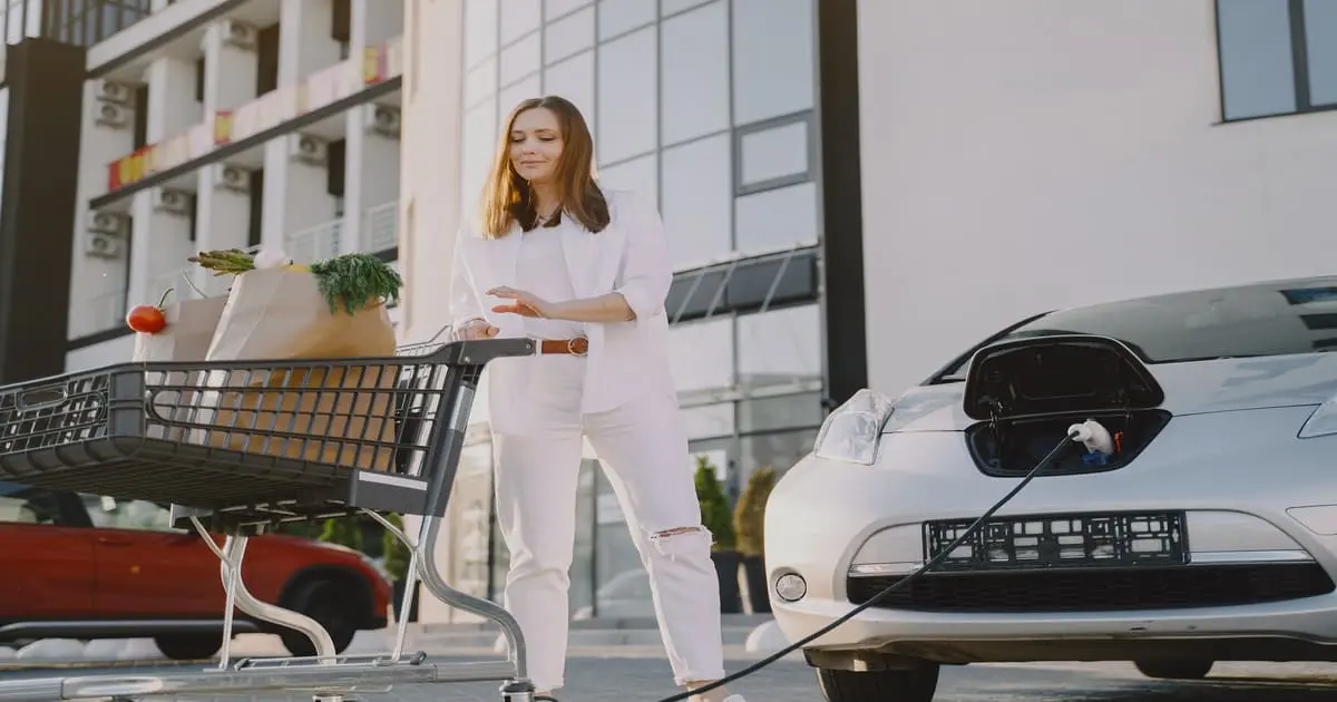 Mulher sorridente, colocou seu carro elétrico para carregar e foi às compras no supermercado. Sustentabilidade e estilo de vida moderno se encontram.