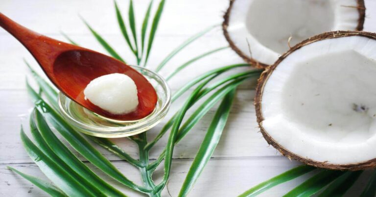 Óleo de coco em uma colher de madeira sobre uma tigela de vidro, rodeado por folhas verdes e pedaços de coco aberto ao fundo.