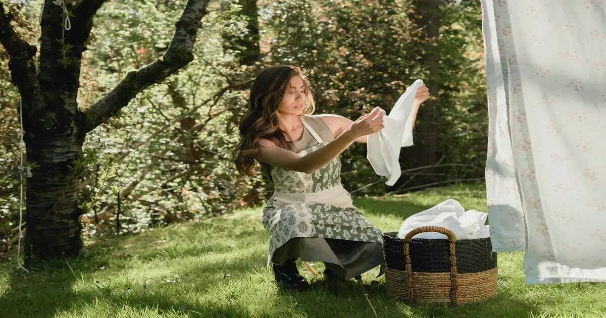 Mulher com avental floral estende um pano ao ar livre, ao lado de um cesto com roupas, em um gramado ensolarado cercado por árvores.