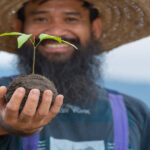 Homem sorridente segurando muda de planta para agrofloresta, simbolizando sustentabilidade, regeneração do solo e cultivo ecológico.