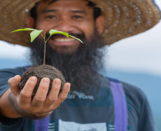 Homem sorridente segurando muda de planta para agrofloresta, simbolizando sustentabilidade, regeneração do solo e cultivo ecológico.