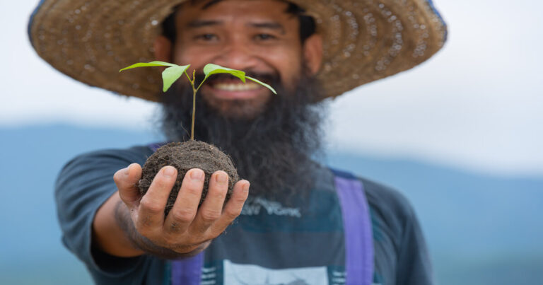 Homem sorridente segurando muda de planta para agrofloresta, simbolizando sustentabilidade, regeneração do solo e cultivo ecológico.