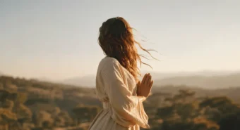 Mulher com as mãos em namaste contemplando o pôr do sol nas montanhas em uma prática de santosha e contentamento interior.